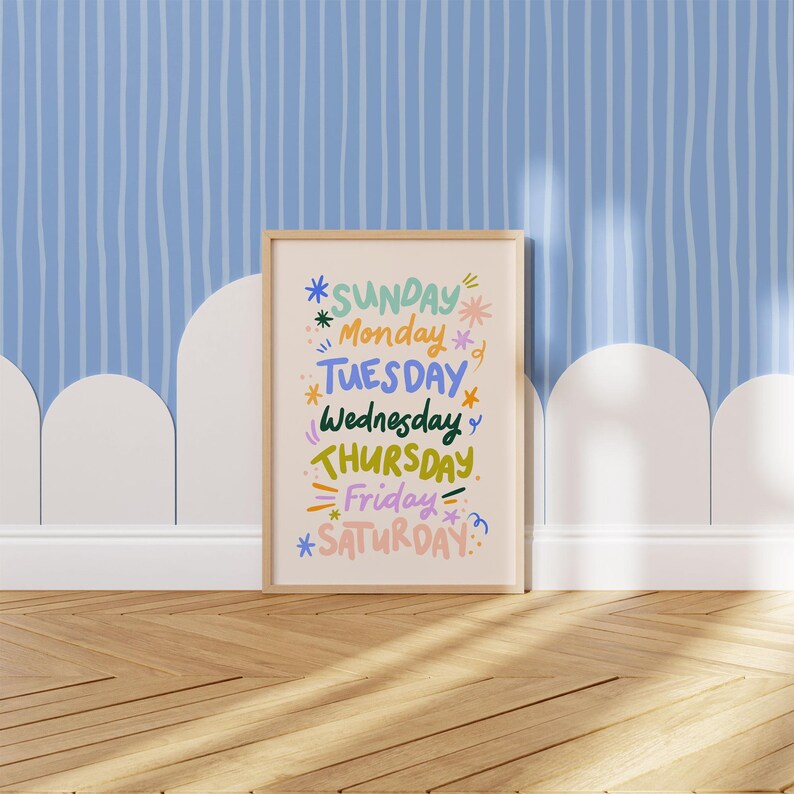 May include: A framed print featuring the days of the week in colorful, whimsical lettering. The days are arranged vertically, with each day highlighted with decorative elements like stars and lines. The frame is a light wood color, and the background is a light beige.