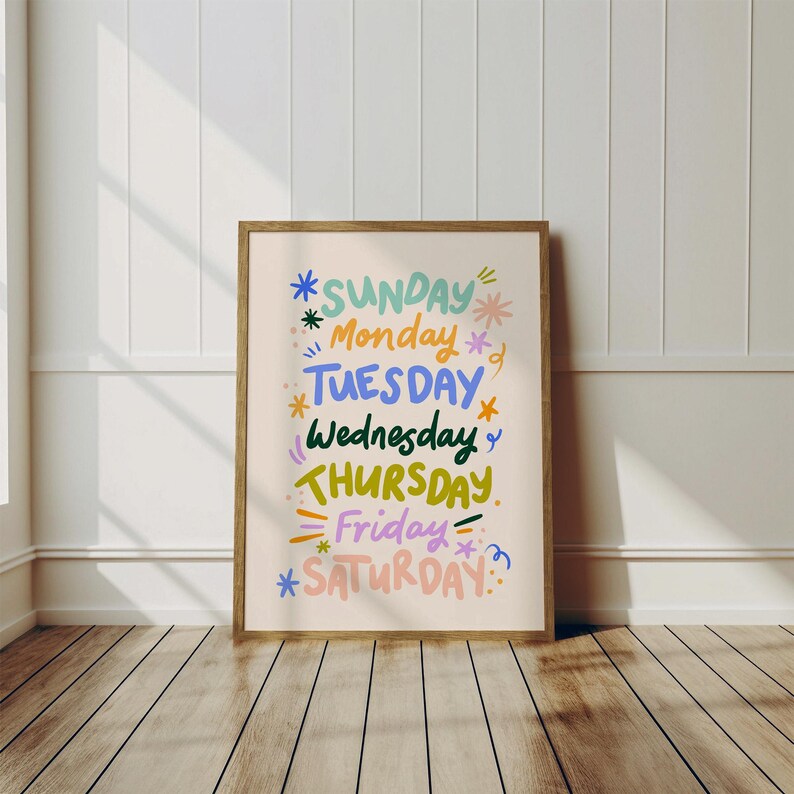 May include: A framed print with the days of the week in colorful, whimsical lettering. The days are arranged vertically, with each day highlighted in a different color, such as blue, green, and pink. The frame is a light brown color.