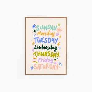 May include: A framed art print featuring the days of the week in colorful, whimsical lettering. Each day is written in a different color, with decorative starbursts and accents. The days are arranged vertically on a cream-colored background.