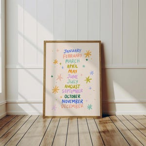 May include: A framed print featuring the months of the year, each month is written in a different color. The print is decorated with colorful starbursts. The frame is a warm brown color, and the background is a light beige.