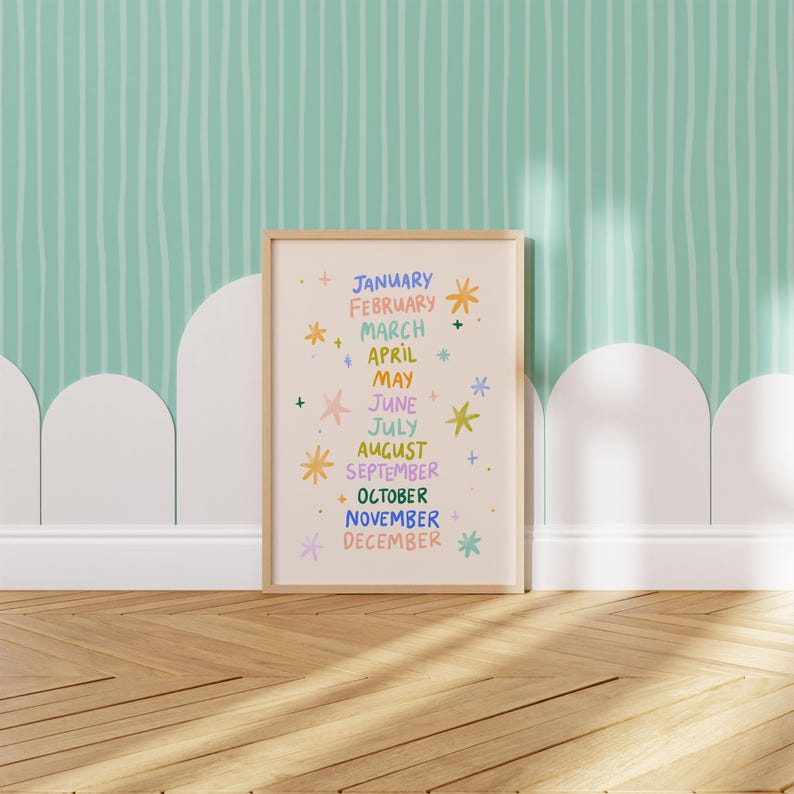 May include: A framed print featuring the months of the year in colorful lettering. Each month is written in a different color, with decorative starbursts and plus signs. The print is in a light wood frame, set against a light green and white striped wall.