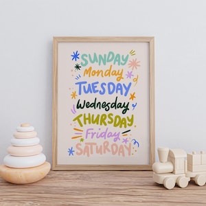 May include: A framed print with the days of the week in colorful, whimsical lettering. The artwork features the days of the week: Sunday, Monday, Tuesday, Wednesday, Thursday, Friday, and Saturday. A wooden toy train and a wooden stacking toy are also in the image.