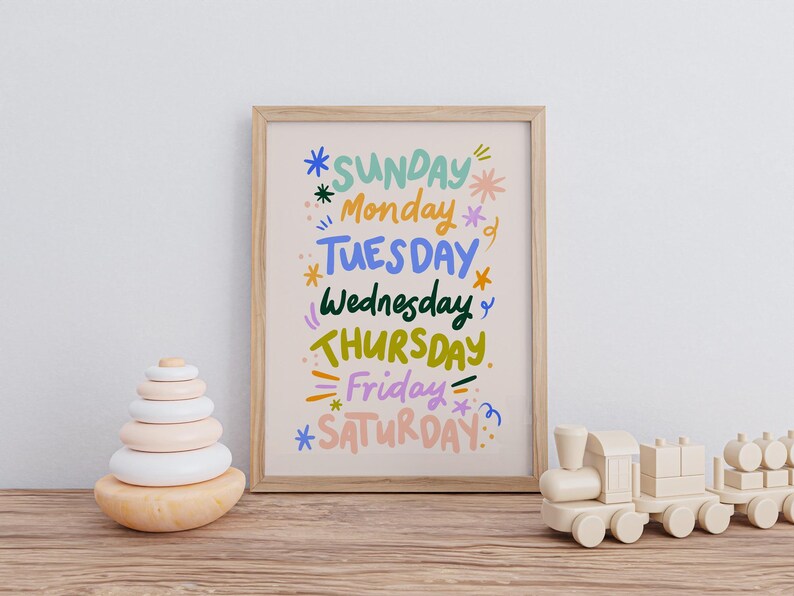 May include: A framed print with the days of the week in colorful, whimsical lettering. The artwork features the days of the week: Sunday, Monday, Tuesday, Wednesday, Thursday, Friday, and Saturday. A wooden toy train and a wooden stacking toy are also in the image.