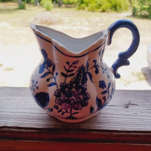 May include: A white ceramic pitcher with a blue handle and floral design. The pitcher has a blue and white floral pattern with a large flower in the center.