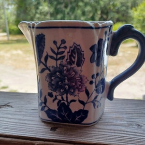 May include: A white ceramic creamer with a blue floral design and a blue handle. The creamer has a square shape with rounded corners.