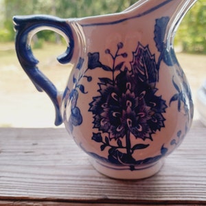 May include: A white ceramic creamer with a blue floral design. The creamer has a handle and a spout.