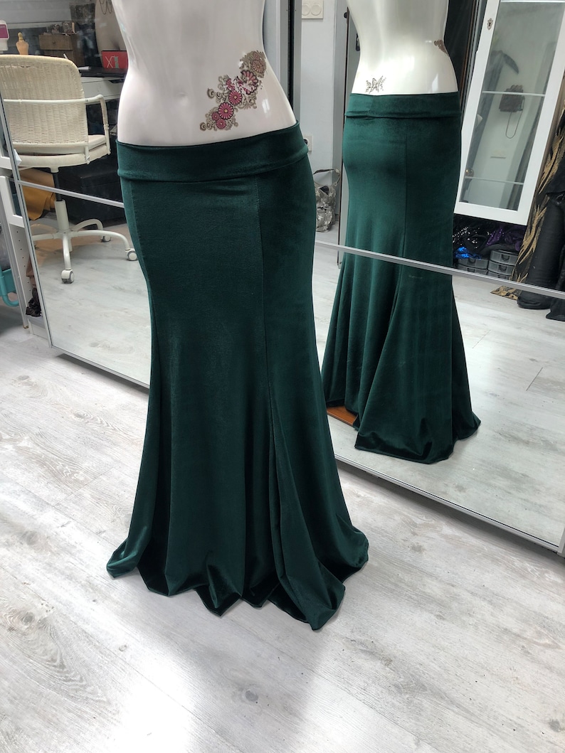 May include: A dark green, floor-length skirt with a mermaid silhouette. The skirt is displayed on a mannequin and has a wide waistband. The fabric appears to be a soft velvet or similar material, with a slight sheen. The skirt flares out from the knees.