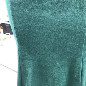 May include: Close-up of a dark teal velvet fabric. The fabric has a soft, textured appearance with subtle variations in color. The image shows the edge of the fabric, highlighting its smooth, flowing lines. The overall impression is one of luxury and depth.