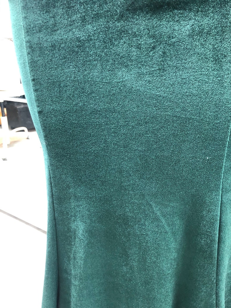 May include: Close-up of a dark teal velvet fabric. The fabric has a soft, textured appearance with subtle variations in color. The image shows the edge of the fabric, highlighting its smooth, flowing lines. The overall impression is one of luxury and depth.