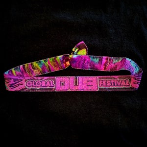 May include: A vibrant pink wristband with the text "GLOBAL DUC FESTIVAL" in a raised, textured font. The band features a colorful, swirling pattern of green, yellow, and purple. The wristband is tied at the top, creating a loop.