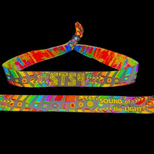May include: A colorful tie-dye wristband with a black plastic clasp. The wristband has a gray background with a repeating pattern of yellow, green, blue, and red arrows and circles. The text "STS9" is printed in yellow on the wristband. The bottom of the wristband has the text "SOUND of the LIGHT" printed in yellow.