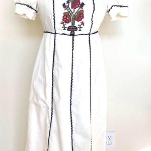 May include: White vintage dress with short sleeves and a square neckline. The dress features a floral embroidered design on the bodice and dark blue trim. The dress is displayed on a mannequin.