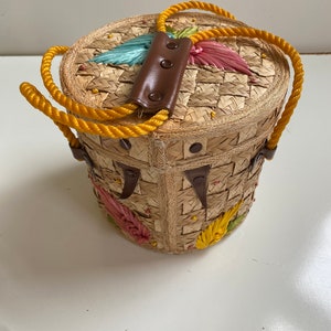 Op de afbeelding: Een ronde strooien tas met een bruin leren handvat en gele touwriemen. De tas is versierd met kleurrijke geborduurde bloemen.