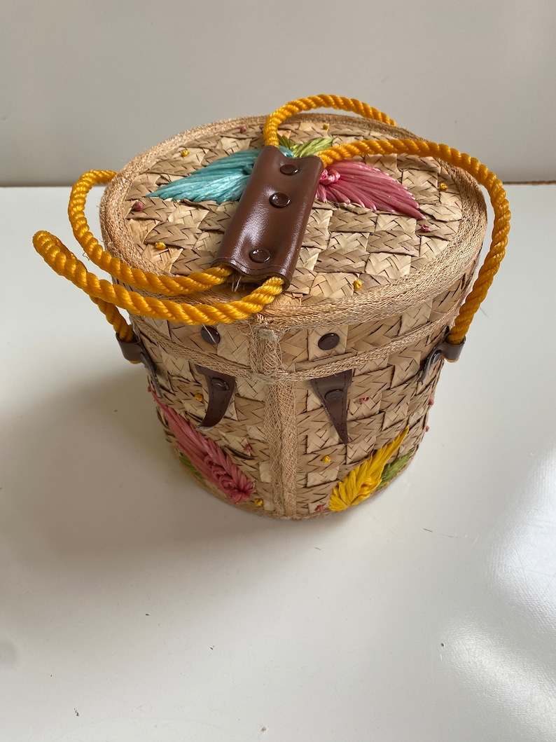 Op de afbeelding: Een ronde strooien tas met een bruin leren handvat en gele touwriemen. De tas is versierd met kleurrijke geborduurde bloemen.