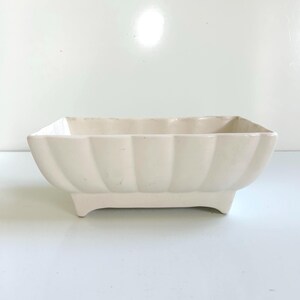 May include: A rectangular off-white ceramic planter with a scalloped, fluted exterior design. The vessel features a smooth, glossy finish and is supported by four short, integrated feet at the base, creating a raised profile for the container.