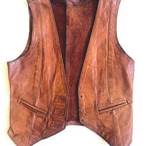 May include: A sleeveless brown leather vest featuring a V-neckline, vertical seam detailing, and two front welt pockets. The garment has a smooth exterior finish and a dark brown interior lining, with a pointed hemline at the front.