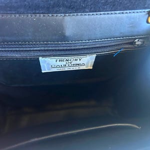 Pode incluir: Bolsa preta com fecho de aba. O interior &eacute; vis&iacute;vel, revelando uma etiqueta que diz "FRENCHY OF CALIFORNIA MADE IN U.S.A.". A bolsa tem um z&iacute;per e um fecho dourado.