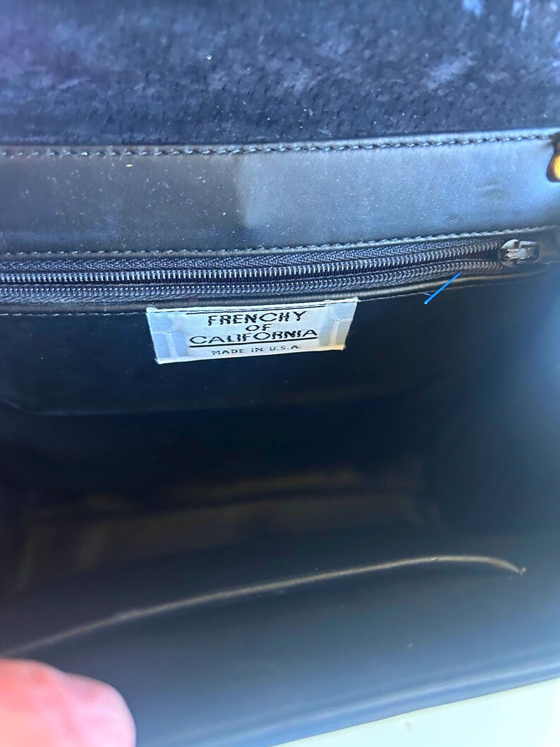 Pode incluir: Bolsa preta com fecho de aba. O interior &eacute; vis&iacute;vel, revelando uma etiqueta que diz "FRENCHY OF CALIFORNIA MADE IN U.S.A.". A bolsa tem um z&iacute;per e um fecho dourado.