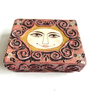 May include: A square, hand-painted trinket box with a sun face design. The box is primarily pink with a yellow center and black swirls. Small white and blue flowers are painted on the corners. The sun face has brown eyes and a small red mouth.