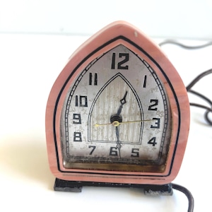 May include: A vintage pink alarm clock with a silver face and black numerals. The clock has a unique arched shape and sits on a black base. The clock's hands are gold-colored, and a black cord extends from the back.