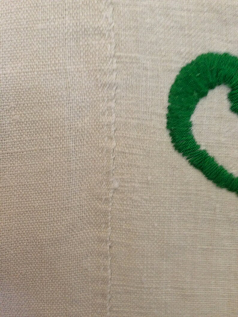 K&ouml;nnte beinhalten: Nahaufnahme eines wei&szlig;en Leinenstoffs mit einem gr&uuml;nen Stickmuster. Die Stickerei ist eine einfache, geschwungene Form.
