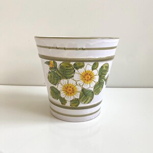 May include: White ceramic planter with a floral design. The planter features a white base with green and yellow floral patterns and green horizontal stripes. The flowers have yellow centers and white petals. The planter is suitable for indoor or outdoor use.