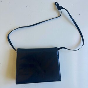 Pode incluir: Uma bolsa transversal de couro preto com formato retangular. A bolsa tem uma al&ccedil;a longa e fina e um fecho de aba. O couro parece liso e brilhante. Um design cl&aacute;ssico e elegante.