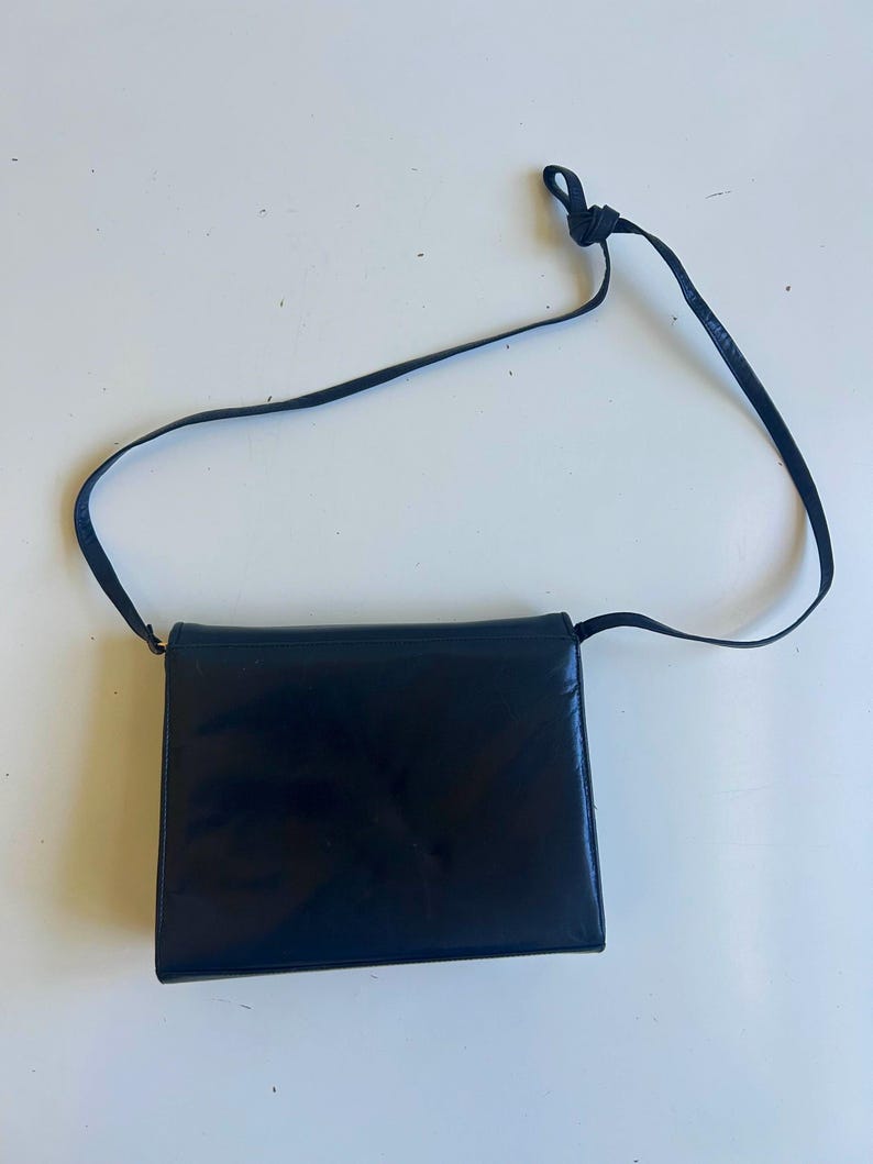 Pode incluir: Uma bolsa transversal de couro preto com formato retangular. A bolsa tem uma al&ccedil;a longa e fina e um fecho de aba. O couro parece liso e brilhante. Um design cl&aacute;ssico e elegante.