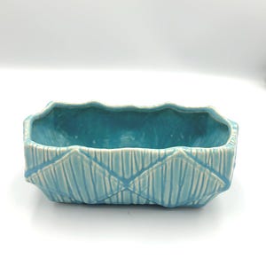 May include: A rectangular, turquoise ceramic planter with a textured, geometric design. The planter has a scalloped rim and is suitable for small plants or succulents. The interior is a darker shade of turquoise.