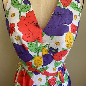 May include: A halter-neck dress featuring a vibrant floral pattern. The dress showcases large, stylized flowers in red, purple, and yellow against a white background. Green leaves and dotted details complete the design. The dress is displayed on a mannequin.