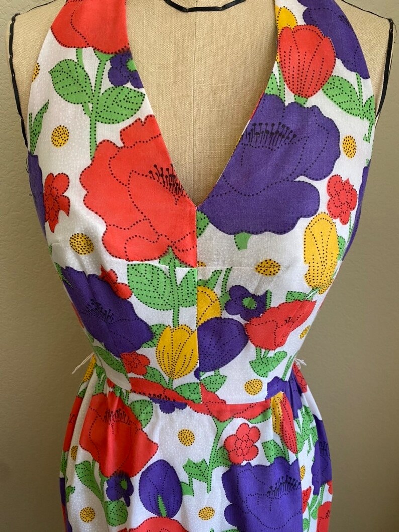 May include: A halter-neck dress featuring a vibrant floral pattern. The dress showcases large, stylized flowers in red, purple, and yellow against a white background. Green leaves and dotted details complete the design. The dress is displayed on a mannequin.