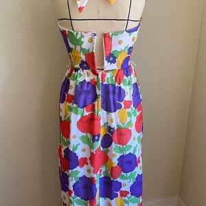 May include: A vintage-style halter dress on a mannequin, featuring a vibrant floral pattern. The dress showcases a white background adorned with large red, purple, and yellow flowers. The halter tie is patterned with the same floral design.
