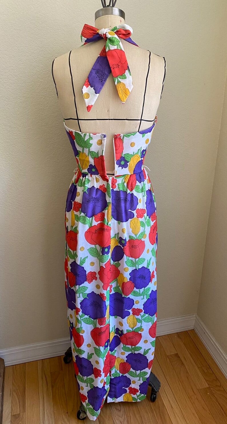 May include: A vintage-style halter dress on a mannequin, featuring a vibrant floral pattern. The dress showcases a white background adorned with large red, purple, and yellow flowers. The halter tie is patterned with the same floral design.
