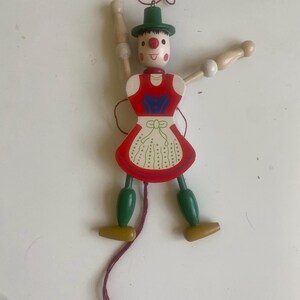 May include: A wooden pull-string marionette doll with a painted face, red apron, and green hat and legs. The doll has jointed arms and legs, and a red string for control. The doll's arms are made of wood and have white spheres at the ends.