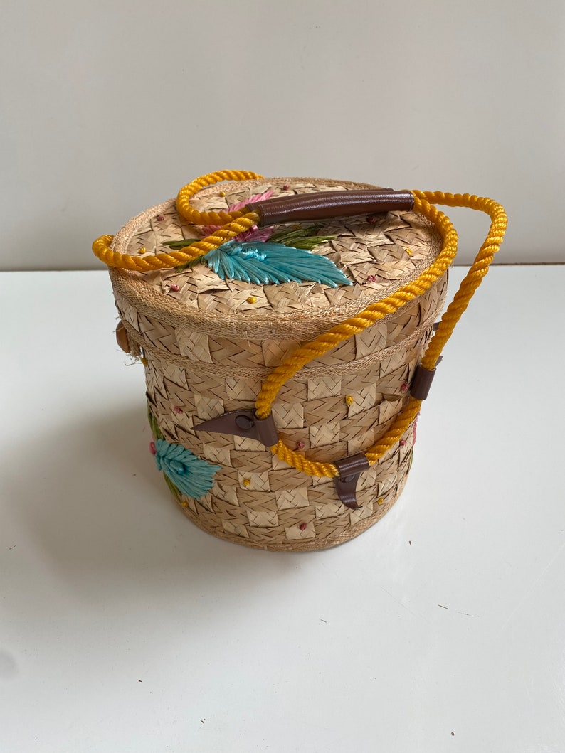 Op de afbeelding: Een ronde geweven strooien mand met een gele touwhandvat en kleurrijke geborduurde bloemen. De mand heeft een bruin leren handvat.