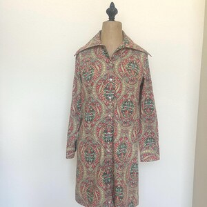 May include: A long-sleeved button-down shirt with a paisley pattern in red, green, and tan. The shirt has a wide collar and is displayed on a mannequin. The fabric has a vintage aesthetic.