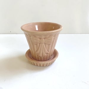 May include: A small, peach-colored ceramic planter with a matching saucer. The planter features a raised bow design on the front and vertical ribbed detailing. The planter is designed for small plants and flowers.