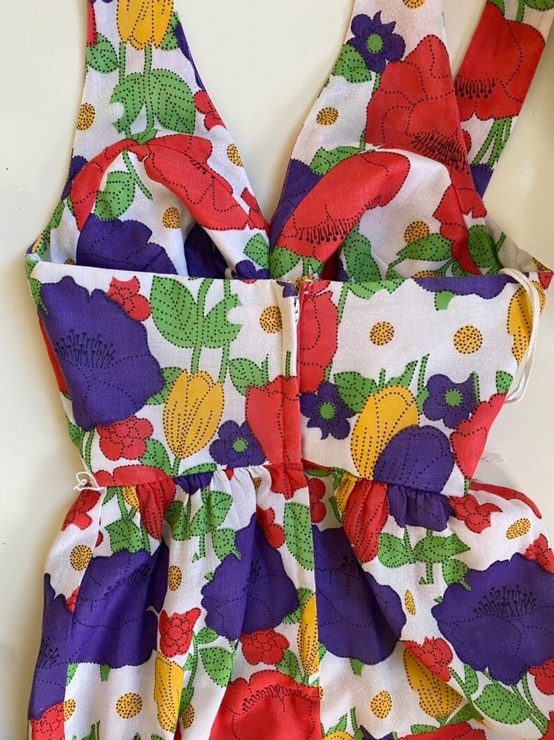 May include: A sleeveless dress with a floral pattern featuring red, purple, and yellow flowers on a white background. The dress has a V-neckline and a gathered waist. The fabric appears to be cotton or a similar lightweight material.