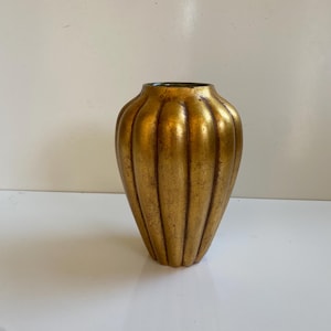 May include: A gold-colored vase with a ribbed design. The vase has a rounded shape with a narrow neck and a wide base. The gold finish has a slightly aged appearance, adding to its vintage charm. The vase is likely made of ceramic or metal.