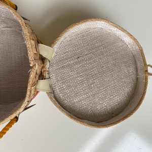 Op de afbeelding: Een ronde, geweven strooien mand met een witte stoffen voering. De mand heeft een gele touwhandvat en een witte leren riem.