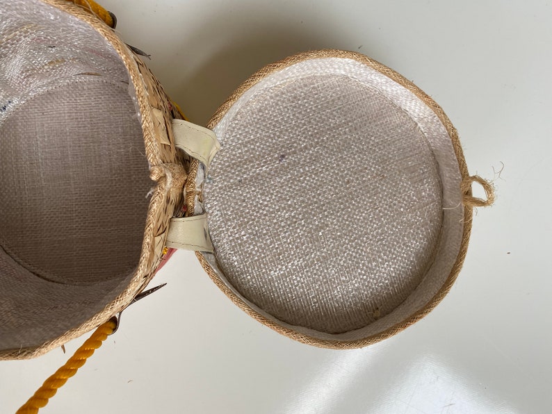 Op de afbeelding: Een ronde, geweven strooien mand met een witte stoffen voering. De mand heeft een gele touwhandvat en een witte leren riem.