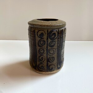 May include: A cylindrical ceramic vase with a brown and green glaze. The vase has a black and white geometric pattern of circles and lines.