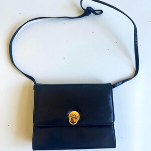 Pode incluir: Bolsa transversal de couro preto com fecho dourado. A bolsa tem formato retangular com aba e uma al&ccedil;a longa e fina. A al&ccedil;a &eacute; amarrada na parte superior. A bolsa est&aacute; em uma superf&iacute;cie branca.