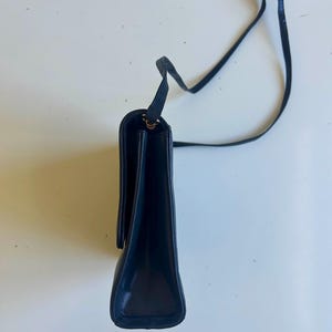 Pode incluir: Uma bolsa de ombro de couro azul marinho com uma al&ccedil;a longa e fina. A bolsa tem formato retangular com fecho de aba. A al&ccedil;a &eacute; amarrada na parte superior. A bolsa est&aacute; em um fundo branco.
