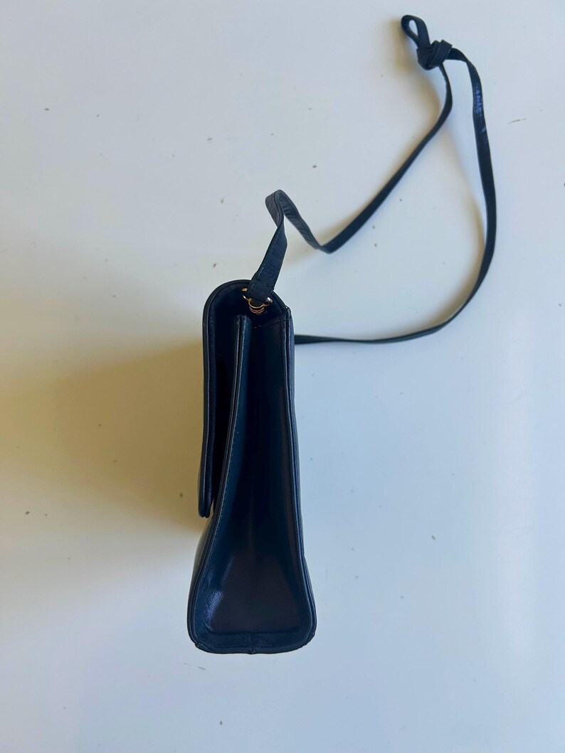 Pode incluir: Uma bolsa de ombro de couro azul marinho com uma al&ccedil;a longa e fina. A bolsa tem formato retangular com fecho de aba. A al&ccedil;a &eacute; amarrada na parte superior. A bolsa est&aacute; em um fundo branco.