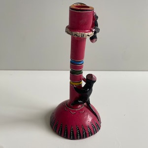 May include: A pink ceramic candle holder with black monkeys climbing up the sides. The holder has a wide base with a black and white geometric design.