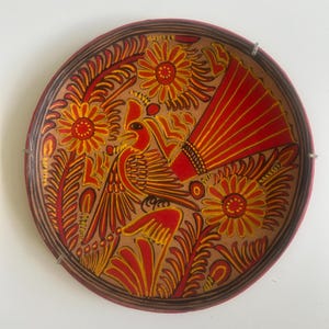 May include: A round ceramic plate featuring a hand-painted design of a bird with a large crest and fanned tail feathers. The artwork uses a palette of red, orange, and yellow against a tan background, surrounded by floral and leaf motifs.