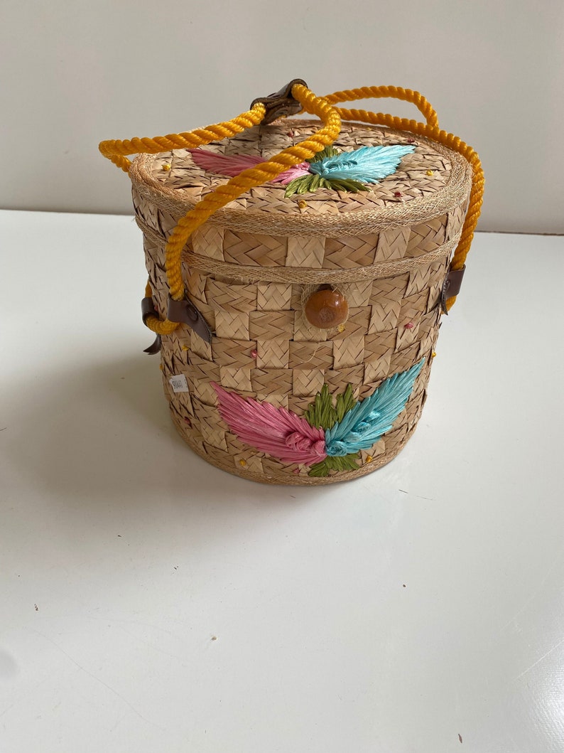 Op de afbeelding: Een ronde geweven strooien tas met een gele touwhandvat en kleurrijke geborduurde bloemen. De tas heeft een geweven deksel en een houten knoopsluiting.