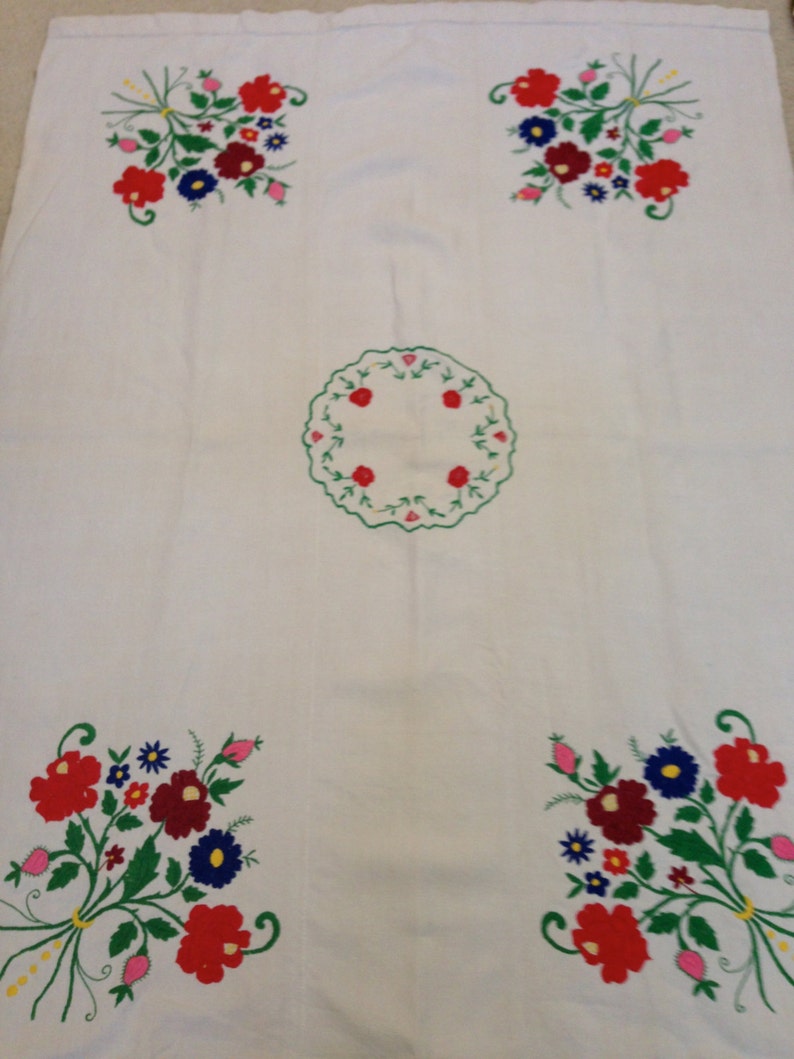 K&ouml;nnte beinhalten: Wei&szlig;e Tischdecke mit gestickten Blumendesigns in Rot, Gr&uuml;n, Blau und Rosa. Die Tischdecke hat ein kreisf&ouml;rmiges Design in der Mitte.