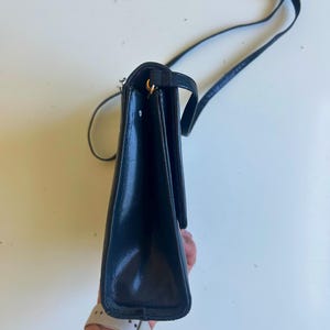 Pode incluir: Bolsa transversal de couro preta com uma al&ccedil;a longa. A bolsa tem formato retangular com fecho de aba e detalhes dourados. A al&ccedil;a &eacute; fina e ajust&aacute;vel. A bolsa &eacute; mostrada de lado.